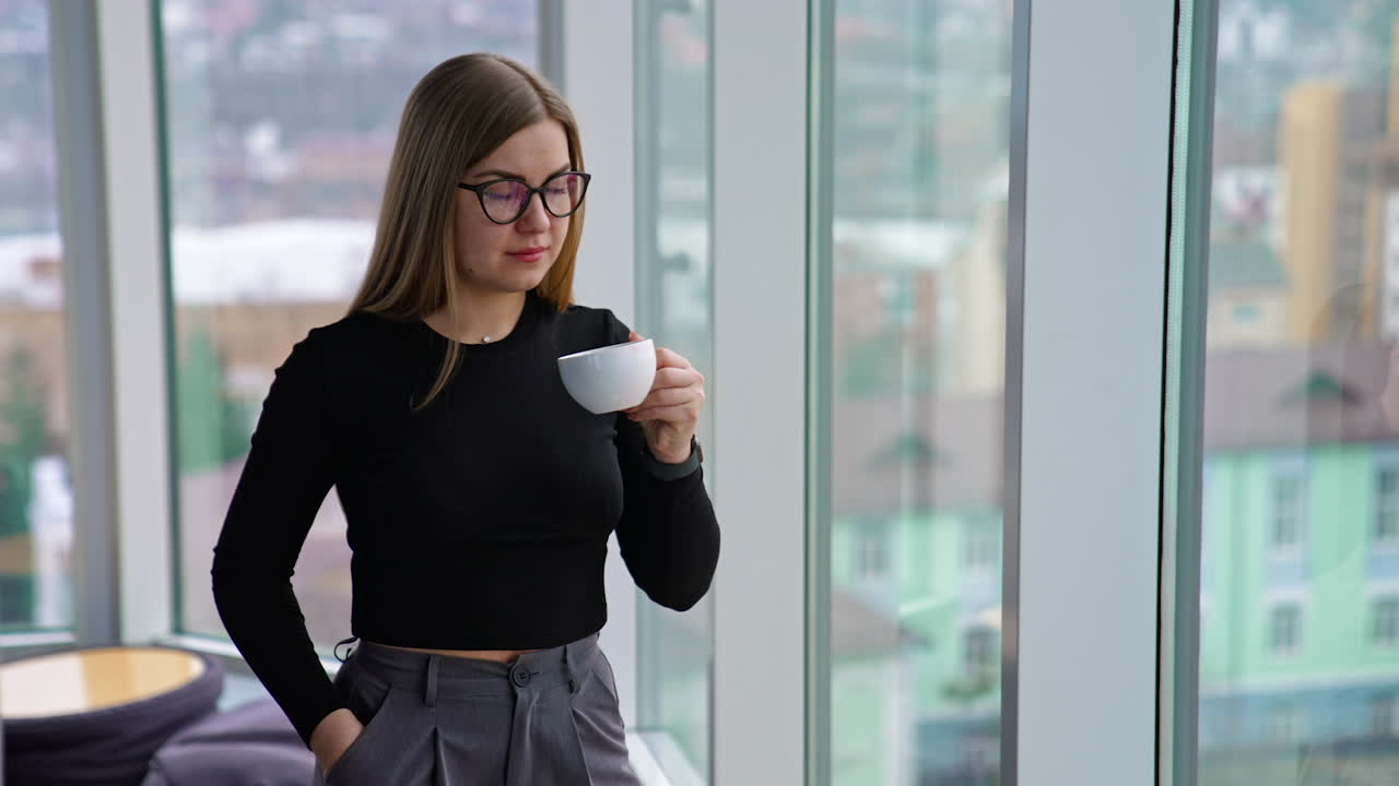 Business lady with a cup of coffee indoors. Young woman in glasses having a break while standing near the window in office and drinking hot beverage.