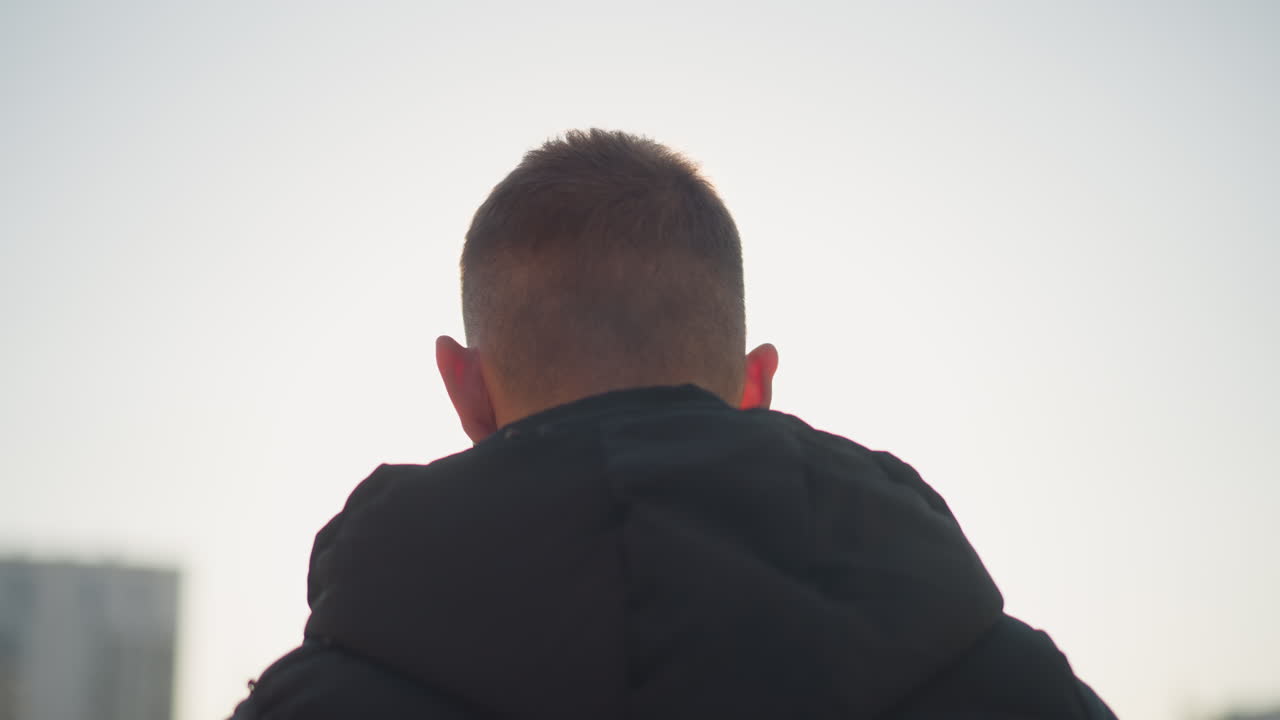 back view of young man in black vest exhaling smoke into sunlight with modern city buildings softly blurred in background creating atmospheric feel of solitude and urban mood