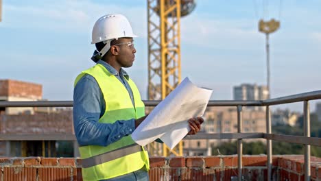 retrato del constructor afroamericano con casco y gafas que sostiene planos y borradores y los estudia mientras está al aire libre en el sitio de construcción.