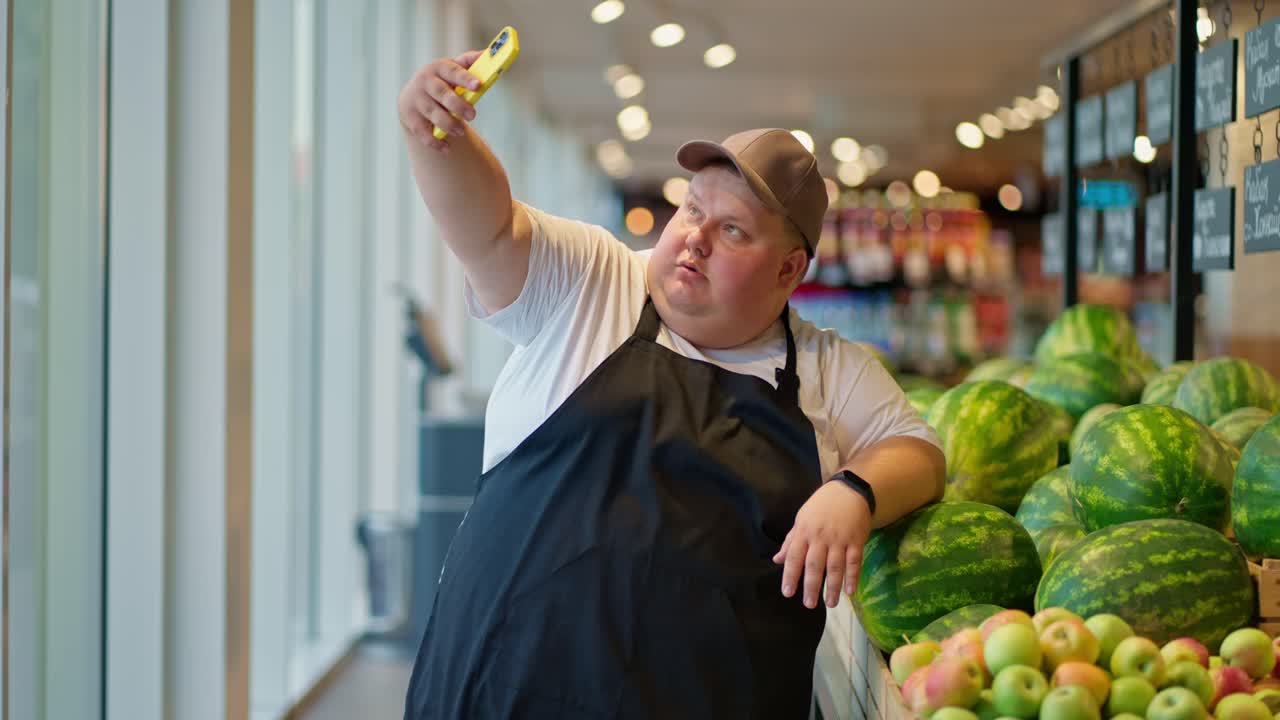 화려하고 행복한 과체중 남성 슈퍼마켓 직원이 색 티셔츠와 회색 모자를 입은 검은색 전신을 입고 슈퍼마크에서 수박을 배경으로 노란색 전화를 사용하여 셀피를 찍습니다.