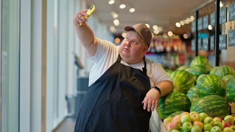 een grappige en gelukkige overgewicht mannelijke supermarktwerker in een wit t-shirt en een zwart schort in een grijze pet neemt een selfie met een gele telefoon tegen de achtergrond van watermeloenen in de supermarkt