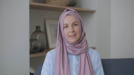 looking at the camera muslim girl calling friends from apartments using a video link waving with hand sitting on a sofa in a stylish room in front of computer screen having video conversation