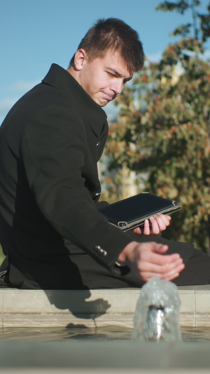 야외 도시 환경에서 초록색, 현대적인 건물, 밝은 빛으로 둘러싸인 태블릿을 사용하는 동안 물 에 앉아서 그 표면을 사려 깊게 만지고 있는 남자