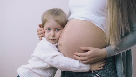 pregnant woman with belly hugs little daughter at light wall