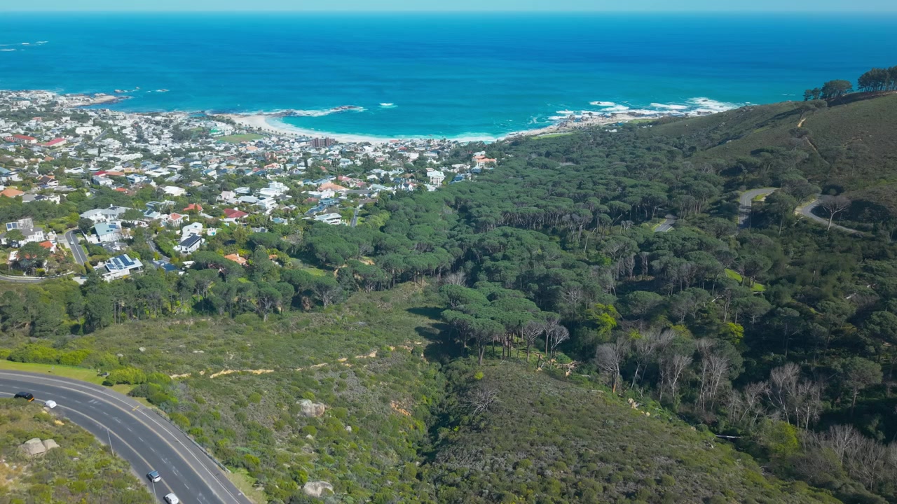 Laaja ilmakuva dronella Camps Bayn ja Bakovenin yllä Kapkaupungin Atlantin rannikolla, Kloof Nek -tien mutkitellessa metsäisten rinteiden läpi kohti alapuolella olevaa rannikkolähiötä.