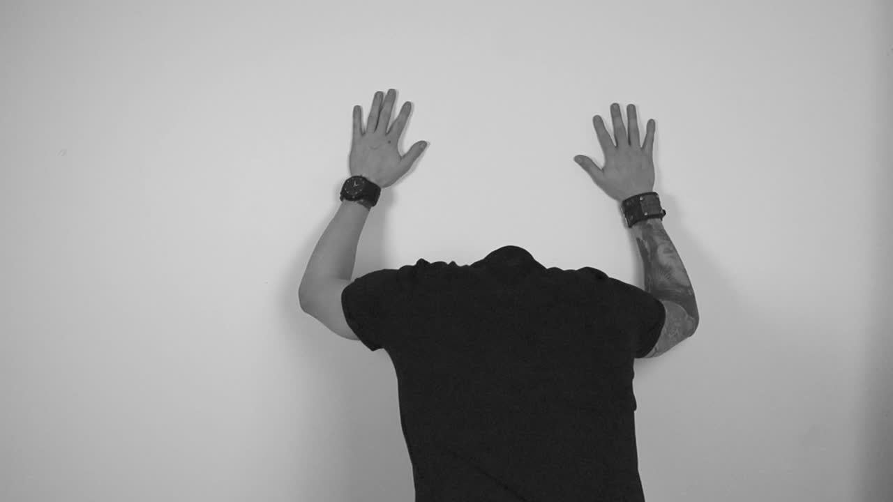 Grayscale footage of a tattooed man expressing anger and frustration by turning to the wall behind him, puts his hands up on the wall and starts pounding and hitting the wall in slow motion.