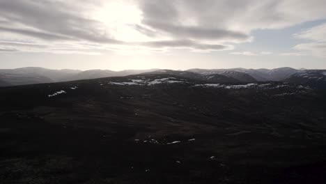 Filmische-Drohnenaufnahmen-Aus-Der-Luft,-Die-über-Einer-Berglandschaft-Und-Heidemoorlandschaft-Mit-Schneeflecken-Aufsteigen,-Während-Die-Sonne-Beginnt,-Hinter-Den-Wolken-Unterzugehen