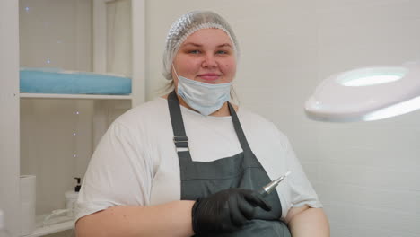 nail technician wearing apron and gloves gently pulls down face mask while holding electric nail file, preparing for session beside lit magnifying lamp in clean salon environment