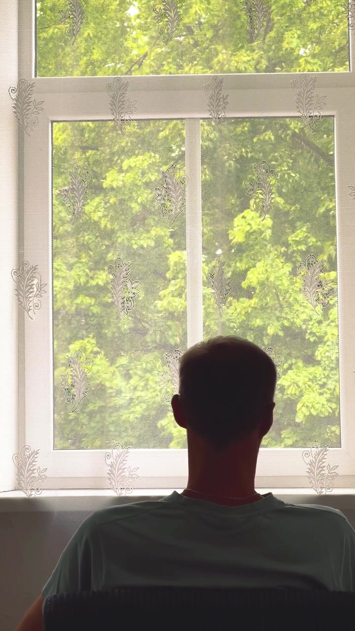 Vertical frame of a man's silhouette looking through a window with lace curtains at bright green summer trees. Ideal for wellness, real estate, or mental health content.