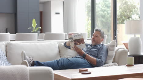 Hombre-Birracial-Senior-Enfocado-Relajándose-En-El-Sofá-Leyendo-Un-Libro-En-La-Sala-De-Estar,-Cámara-Lenta