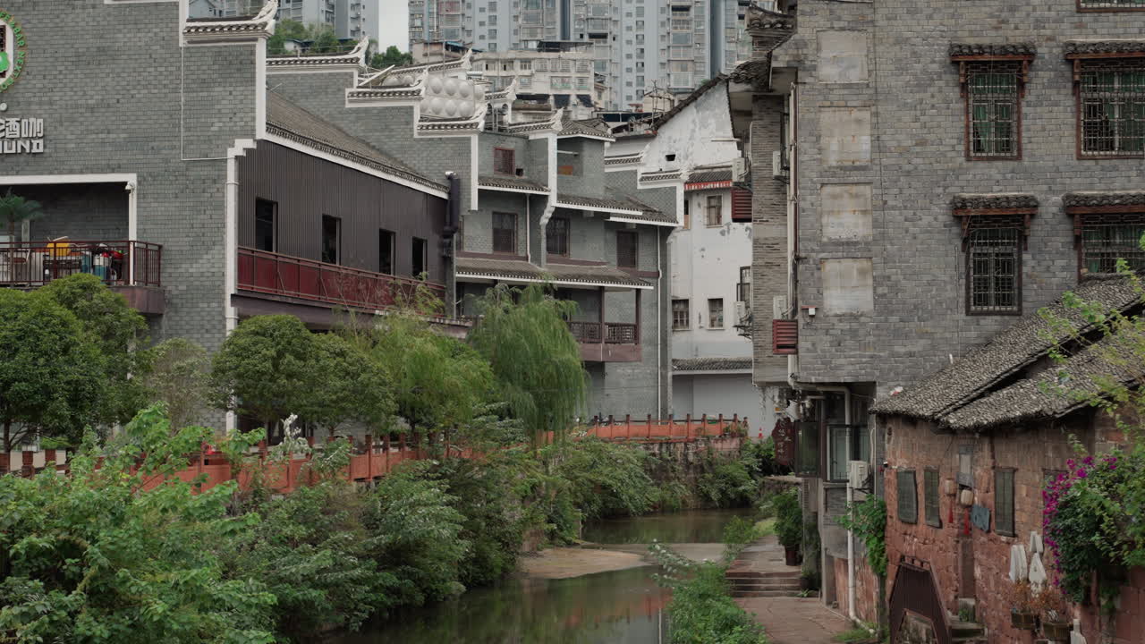 Vicolo fluviale invaso dalla vegetazione tra vecchi edifici, stretto canale che si snoda tra facciate di mattoni, balconi arrugginiti e tetti di tegole, fitta vegetazione che si riappropria del cortile e del sentiero, cielo nuvoloso e cupo