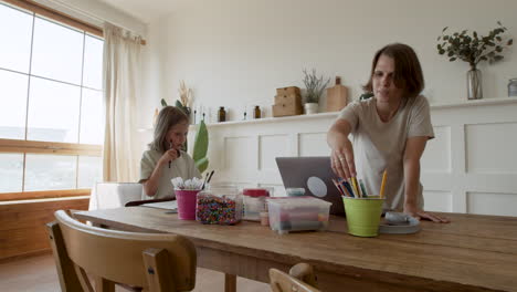 una madre le da lápices a su hija para que pueda dibujar mientras se prepara para trabajar desde casa en su computadora portátil