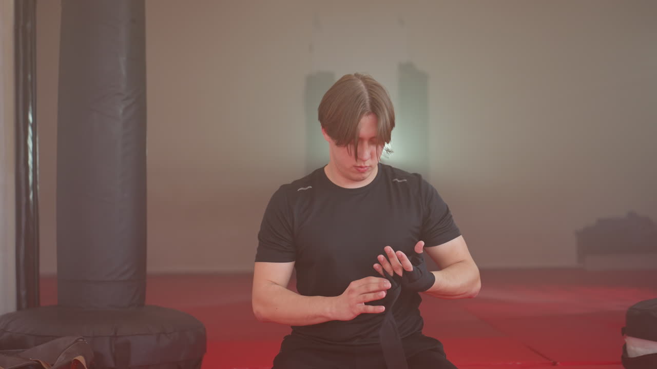 Boxer in black shirt preparing for training, sitting with head down and focused expression while adjusting hand wraps, gym environment with red mats in background showing concentration and readiness
