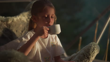 child drinks hot chocolate sitting in furry chair. cute little girl with ponytail closes eyes enjoying pleasant taste of sweet drink at home