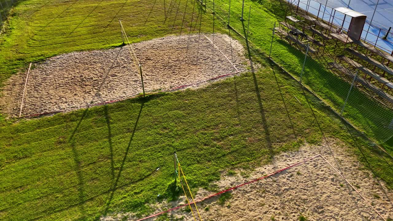 Aerial view of multiple sand volleyball courts surrounded by green fields, captured in warm evening light