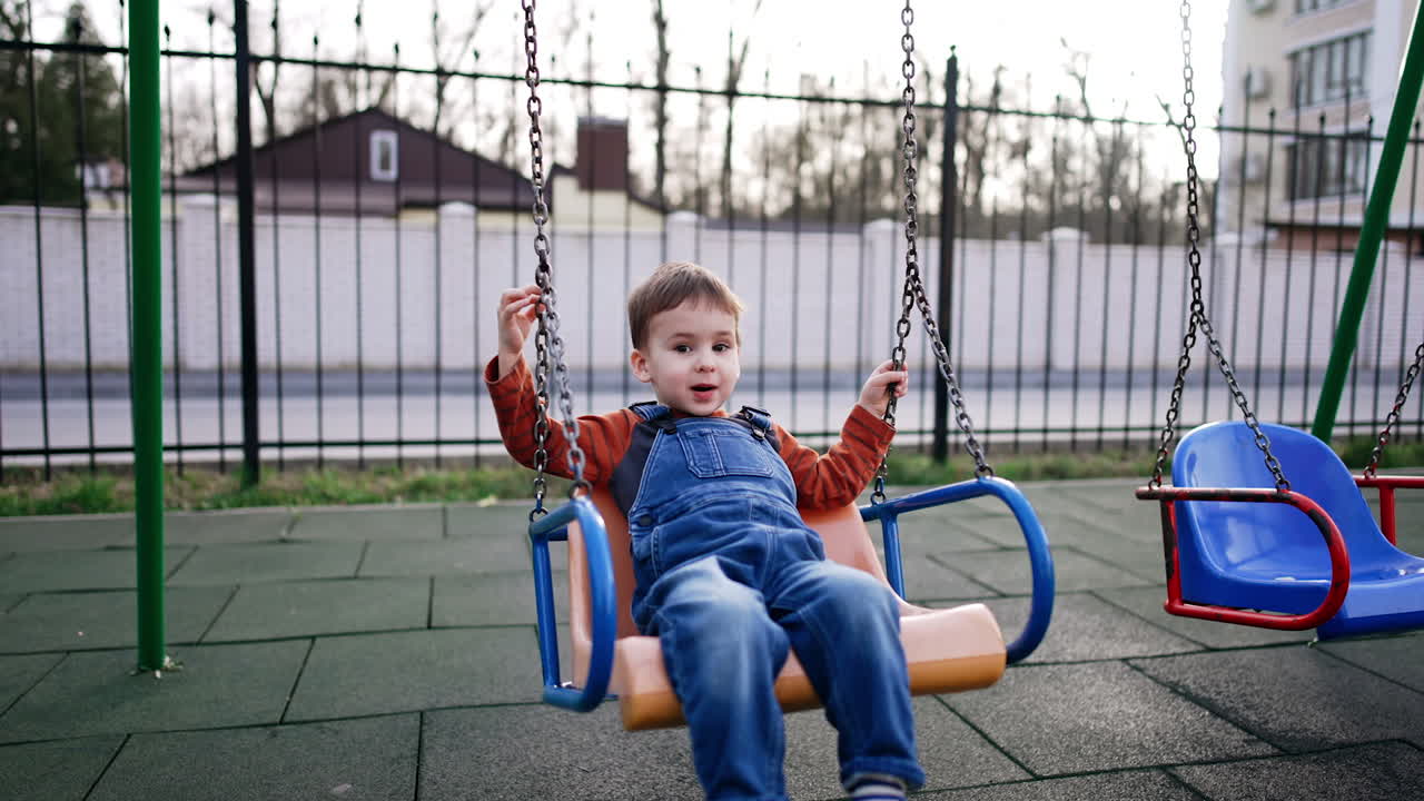 Positive Caucasian toddler swinging at the playground. Baby boy having fun outdoors in autumn.