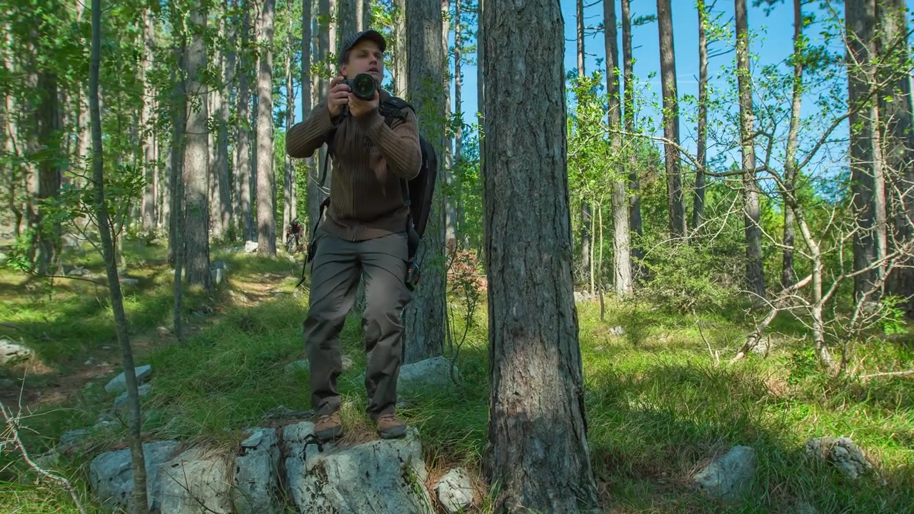 Dotato di macchina fotografica e zaino, un fotografo si trova su una roccia in una foresta, fotografando un'azione sportiva o della fauna selvatica