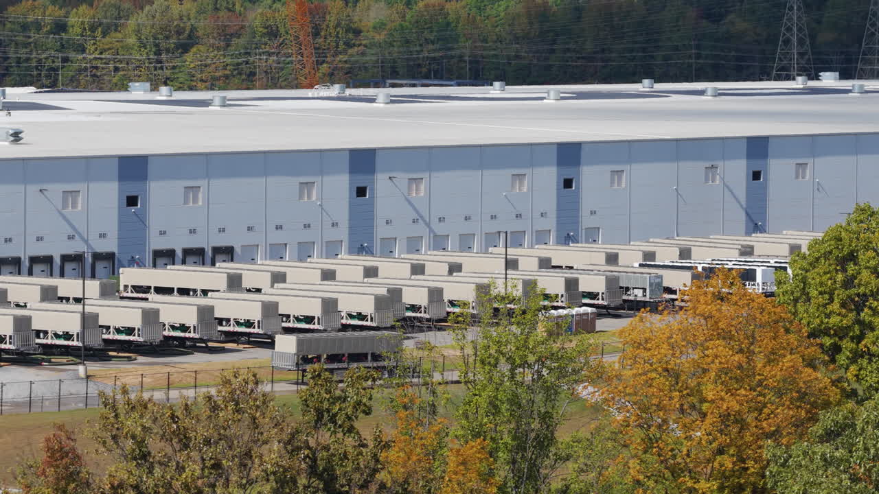 Telephoto drone shot sliding right over autumn trees toward parked mobile power equipment beside a large data facility in Southaven Mississippi United States