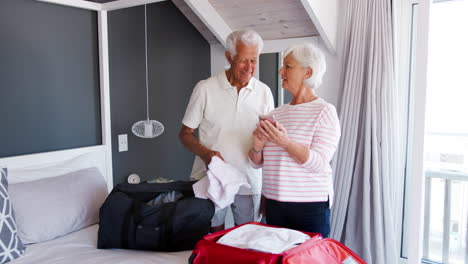 Una-Pareja-De-Ancianos-En-El-Dormitorio-Mira-El-Móvil-Mientras-Empacan-Maletas-Para-Las-Vacaciones