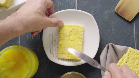 man's hand butters with ghee a soda cracker with a knife on a plate