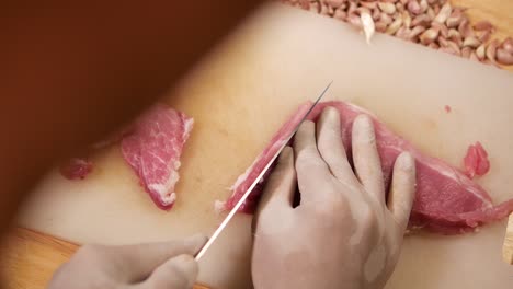 hand with glove use knife to cut pork loin into pieces on white chopping board, close up-1