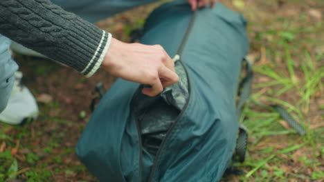 close up of person wearing sweater zipping up green camping bag filled with tent fabric while kneeling on grassy forest floor, surrounded by dry leaves and pine needles during outdoor adventure