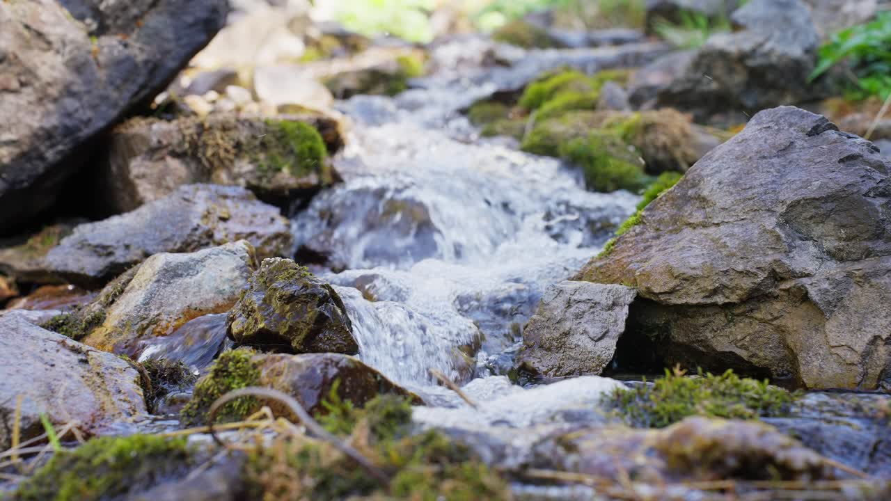 Small mountain stream flows over mossy rocks. Water splashes as it tumbles down stones. Green moss covers wet surfaces. Scene feels calm, peaceful, natural. Great for nature, travel