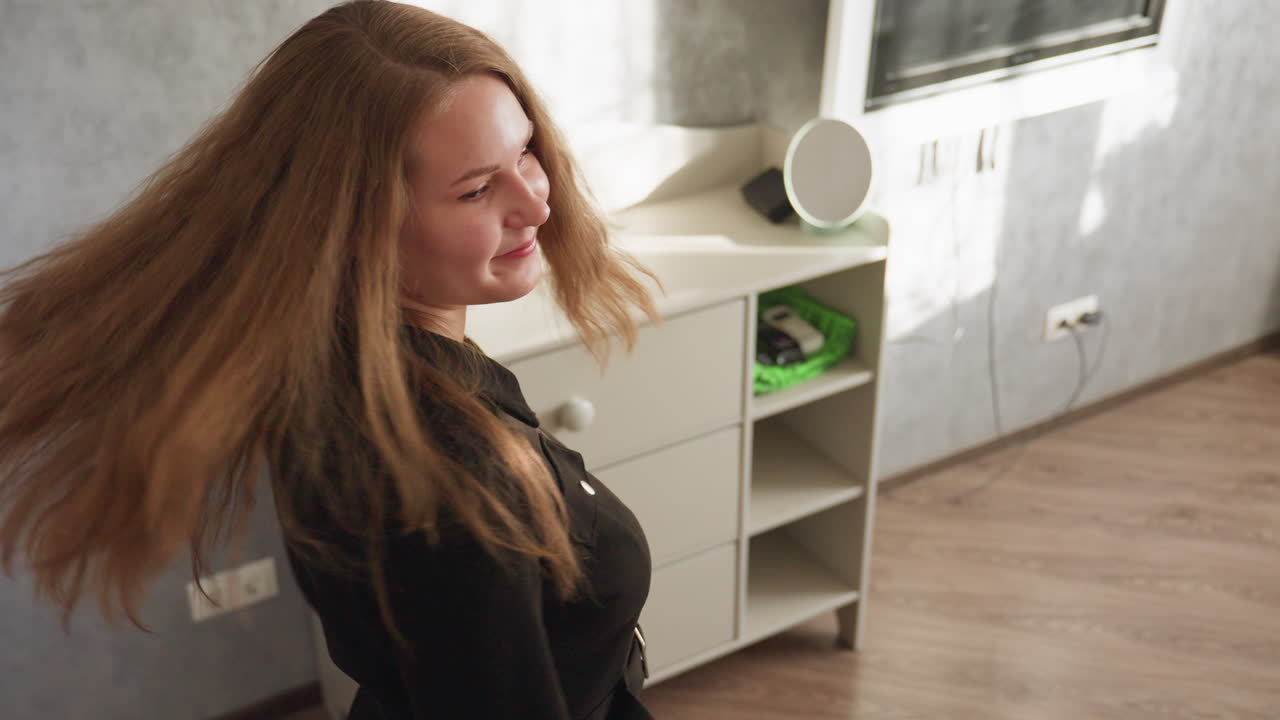 검은색 드레스를 입은 젊은 여성이 우아하게 방으로 걸어 들어가 자연스러운 긴 머리를 자신감 넘치는 자세로 휘날리는 모습이 공중에서 보이는 장면이에요. 부드러운 햇빛이 여성의 움직임에 따라 우아함과 매력을 강조하며 특징을 부각해요.