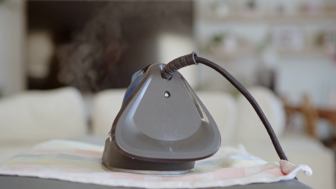 Close up shot of a man's hand using a hot, steaming iron to press a colorful cloth on an ironing board, showing a routine domestic chore in a home environment with a blurred background