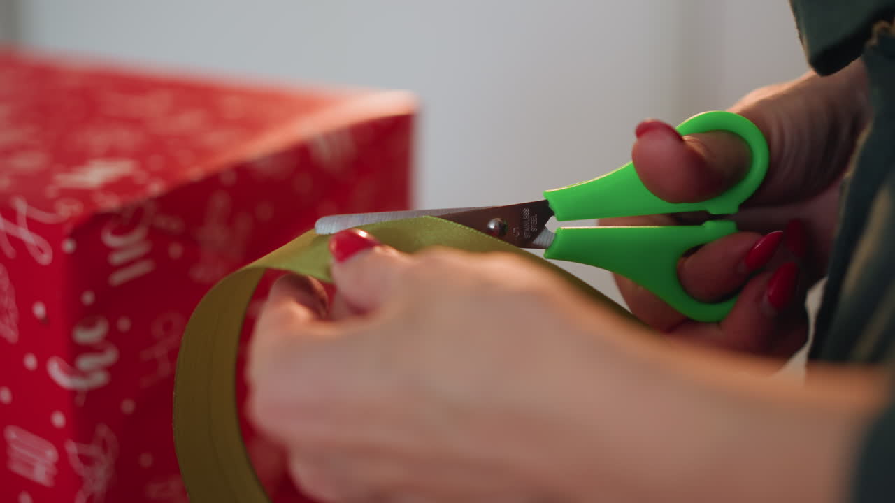 Closeup hands on christmas present ribbon, green scissors snipping gold tape against red patterned paper, wellmanicured nails, intimate indoor holiday atmosphere, meticulous giftwrap preparation