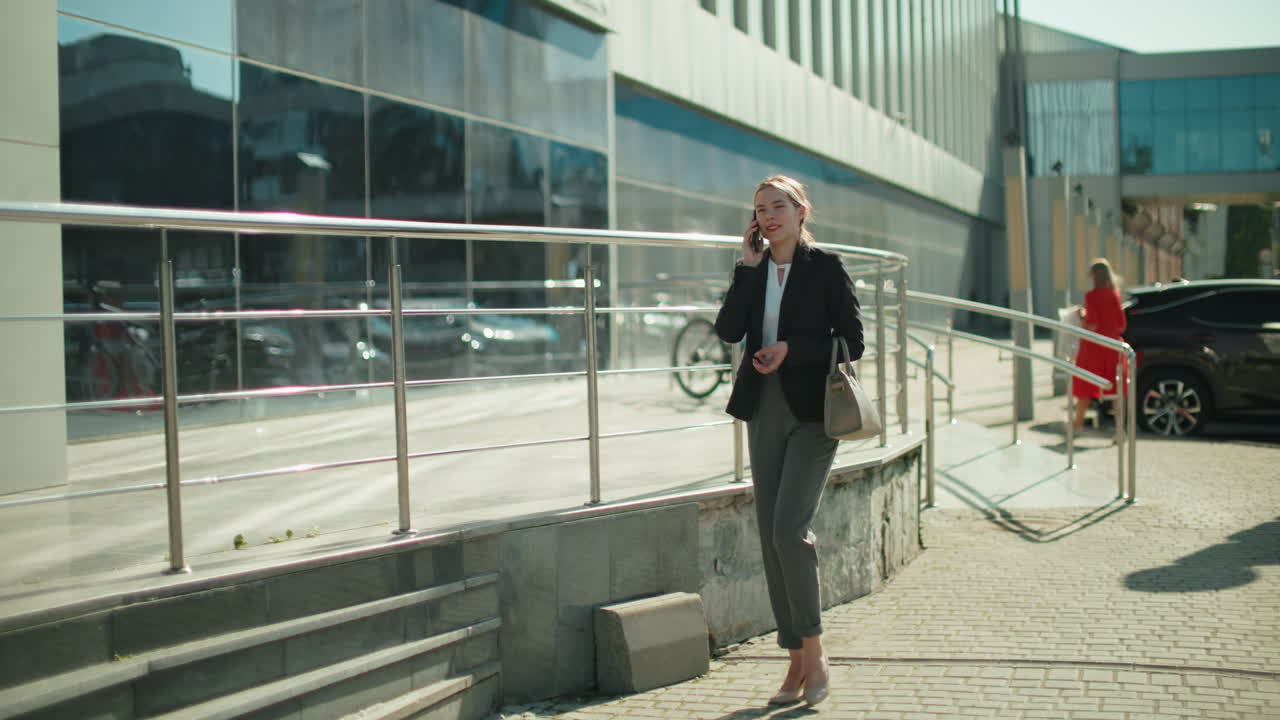Professional business woman walks home while talking on phone, dressed in formal outfit, holding handbag, with urban glass building reflecting surroundings