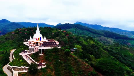a sri lankan hilltop temple with a striking white stupa stands among tropical hills, winding stairways, scattered village houses, and expansive panoramic views.