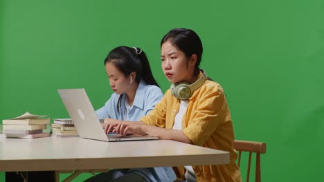 unsatisfied asian woman student sitting with her friend and typing on a laptop on the table in the green screen background classroom