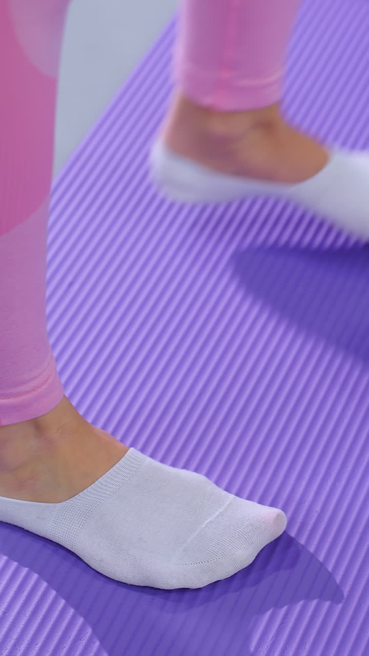 운동 중에 보라색 매트에 균형을 잡고 있는 분홍색 옷을 입은 여성의 다리 시각, 그녀는 잘 조명된 체육관에서 안정성과 힘 훈련에 초점을 맞추는 동안  양말을 입고 있습니다.