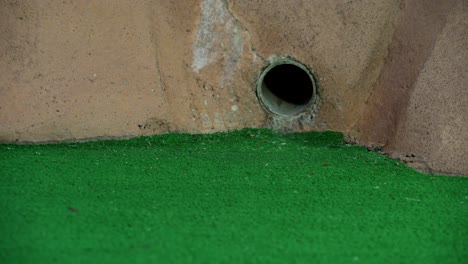 an orange mini golf ball exits a pipe in a wall on a golf course