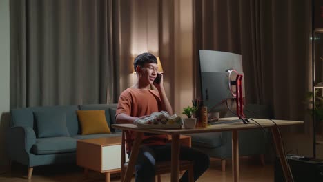 asian boyfootwear designer talking on smartphone while working on a desktop computer at home