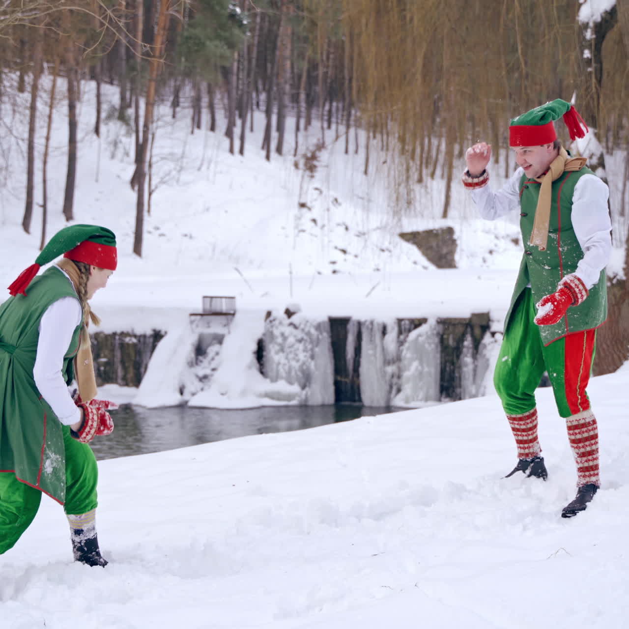 Two funny elves in green costumes playing happily on winter background. One dwarf falling into white snow near the river. Happy Christmas holidays.