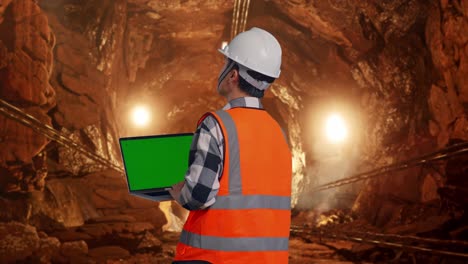 back view of asian male engineer with safety helmet working on a green screen laptop and looking around while standing in underground mine tunnel