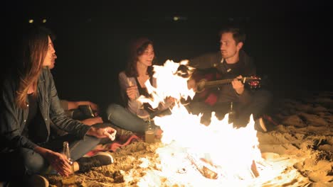 Die-Kamera-Bewegt-Sich-Spät-In-Der-Nacht-Um-Das-Lagerfeuer:-Junge-Fröhliche-Menschen-Sitzen-Abends-Am-Feuer,-Spielen-Gitarre-Und-Trinken-Bier.-Fröhliche-Freunde,-Die-Lieder-Singen,-Reden-Und-Spaß-Haben