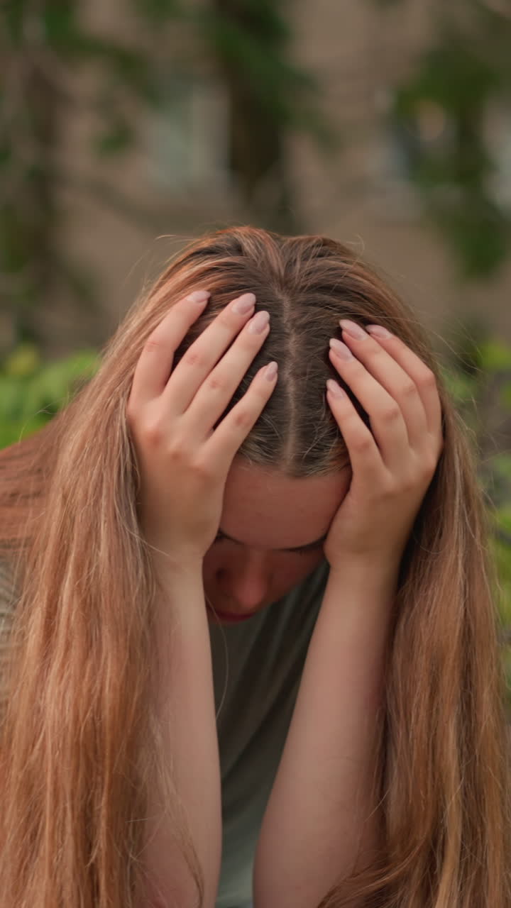 나무 벤치에 앉아있는 여성의 클로즈업, 손으로 얼굴을 고, 반사적이고 우울한 자세로, 그녀의 긴 머리카락이 어깨 아래로 떨어지고, 잔잔한 초록색과 나무가 배경입니다.
