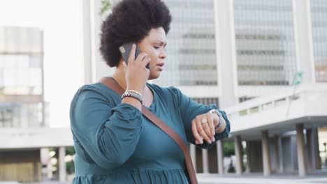 Happy-plus-size-biracial-woman-talking-on-smartphone-and-checking-smartwatch