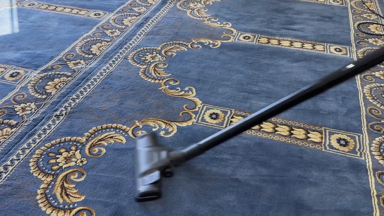 İç Mekanda Desenli Mavi Halı Üzerinde Hareket Eden Elektrik Süpürgesi Başlığı, Rutin Ev Zemin Bakımı, Toz Temizliği ve Ev Hijyenini Gösteriyor