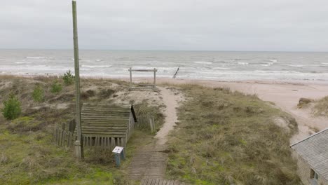 establishing aerial view of baltic sea coast on a overcast day, old wooden pier, white sand beach, large storm waves crushing against the coast, climate changes, wide drone shot moving forward