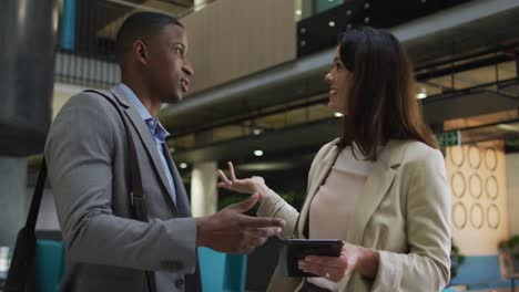 Diverse-businessman-and-businesswoman-talking-and-using-tablet-in-lobby-of-modern-office