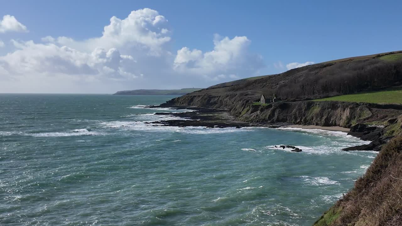 la escarpada costa de irlanda en un día soleado de mayo con olas y campos verdes, zoomando en una lejana iglesia ruinas y tumbas