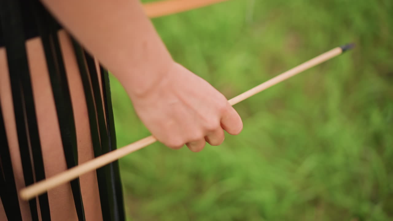 외로운 전사가 나무 지팡이를 쥐고 있어요. 손과 가죽 프린지 스커트를 입은 허벅지를 클로즈업했고, 햇빛이 비치는 풀 배경이에요. 미묘한 움직임은 균형 잡힌 자세를 암시하며, 긴장한 손가락과 보이는 핏줄, 튼튼한 체격을 묘사합니다.