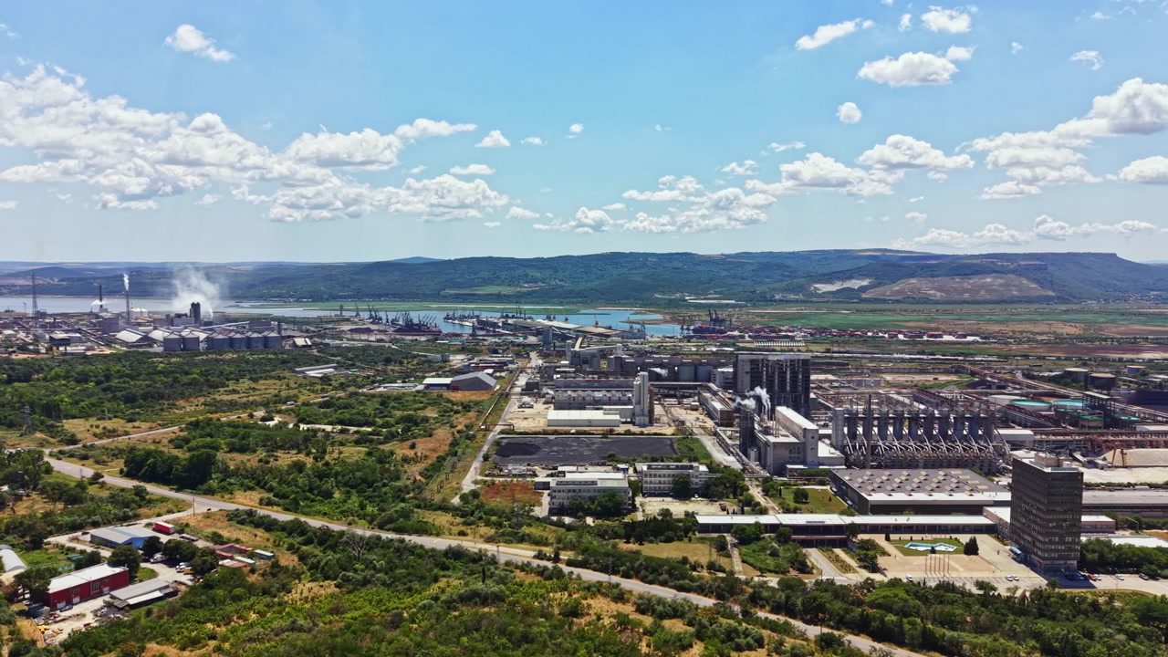 Vista urbana di una vasta area industriale con fabbriche e impianti lungo un fiume, sotto un cielo blu con nuvole.