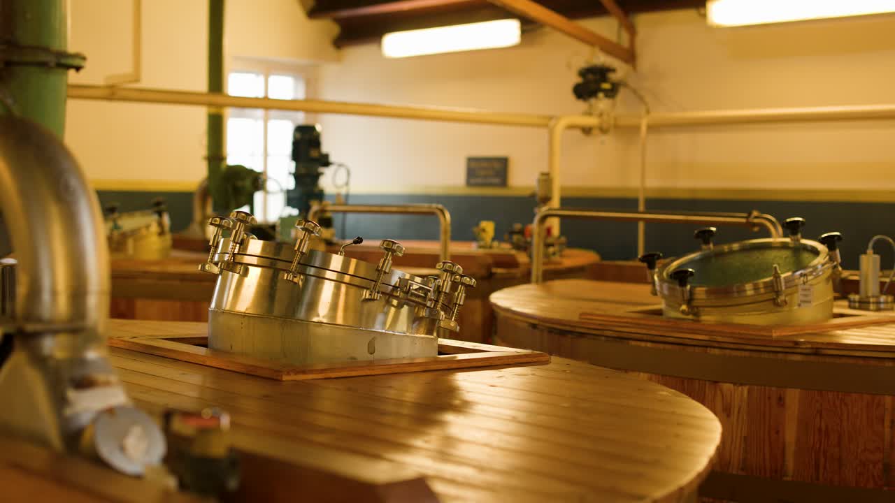 Warmly lit distillery fermentation room with wooden washbacks, metal hatches, and static camera view