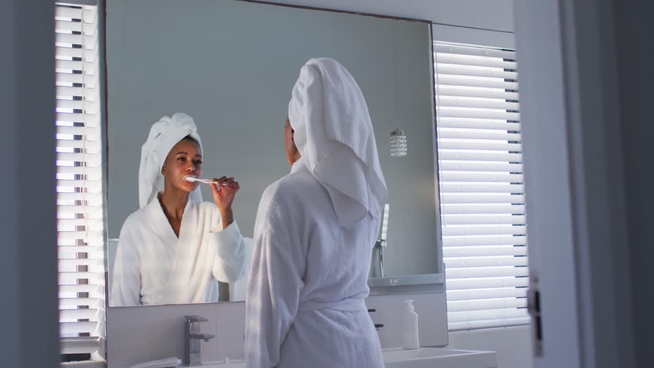 African american woman in bathrobe brushing her teeth looking in the mirror at bathroom