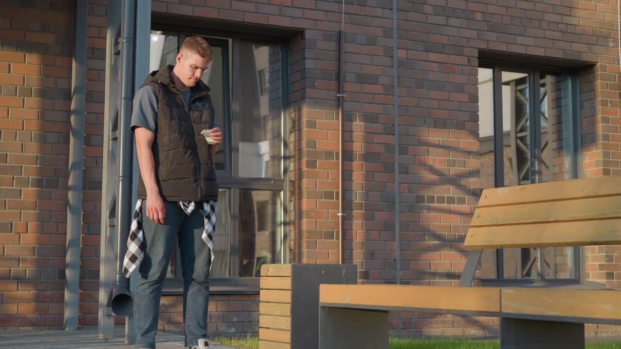 young man dressed in black padded vest and jeans stands beside wooden bench near brick building, looking down as he tosses cigarette pack into waste bin under warm natural sunlight